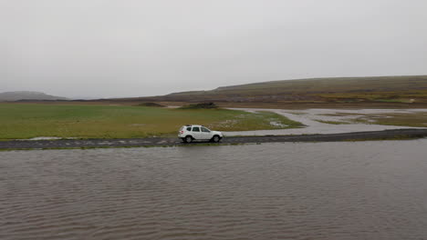 Aerial:-Side-follow-of-white-car-passing-through-a-shallow-river