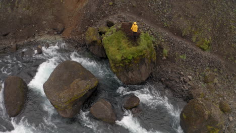Aerial:-Top-down-and-reveal-shot-of-a-man-standing-on-a-rock-on-the-river-bank