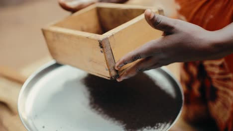 Eine-Afrikanische-Dame-Verwendet-Ein-Handgefertigtes-Sieb,-Um-Dunkelbraun-Gerösteten-Ugandischen-Kaffee-Zu-Sieben