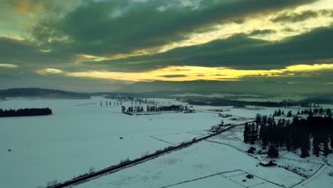 Aerial-Shot-of-Farmland-under-a-Stunning-Yellow-Sky-surrounded-by-snow-covered-grassland-and-needle-forests