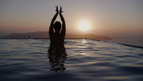 Eine-Frau-Tanzt-Im-Infinity-Pool-Und-Beobachtet-Den-Sonnenuntergang-über-Dem-Meer---Zeitlupe,-Santorini,-Griechenland