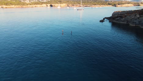 Aerial-View-Behind-Two-People-Surfing-Across-Waters-Off-Cala-Escondida-Coastline-On-Electric-Surfboards-Tracking-Shot
