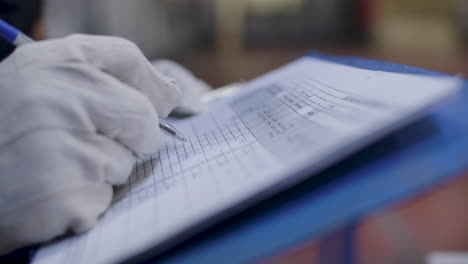 Worker's-Hand-In-Gloves-Writing-On-Monitoring-Sheet-At-Workplace