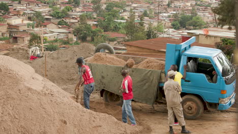 Zwei-Männer-Schaufeln-Sand-In-Einen-Lastwagen-In-Ruanda,-Im-Hintergrund-Häuser