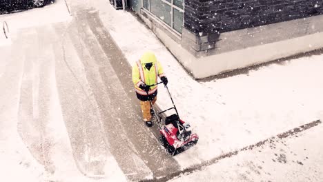 Person-with-reflective-yellow-jacket-plow-snow-of-commercial-building-during-snowfall