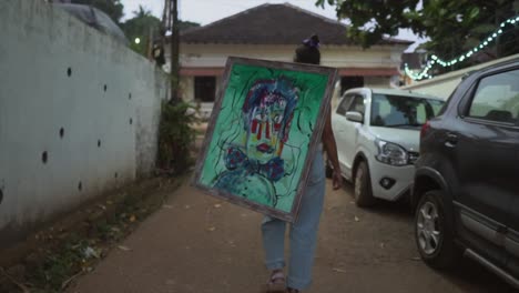 A-shot-following-behind-an-Asian-female-walking-up-a-road-carrying-a-large-picture-frame-with-an-abstract-portrait-painting,-Goa,-India