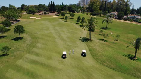 Vogelperspektive-Auf-Einen-Der-Kurzen-Grünen-Rasenflächen-Eines-Golfplatzes,-Auf-Dem-Zwei-Golfwagen-In-Der-Nähe-Von-Golfspielern-Geparkt-Sind