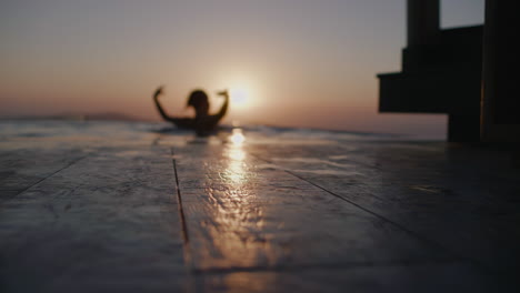 Eine-Frauensilhouette-Tanzt-Im-Infinity-Pool-Bei-Sonnenuntergang-Mit-Sonnenreflexionen-Auf-Dem-Holzboden---Defokussiert,-Zeitlupe,-Santorini,-Griechenland