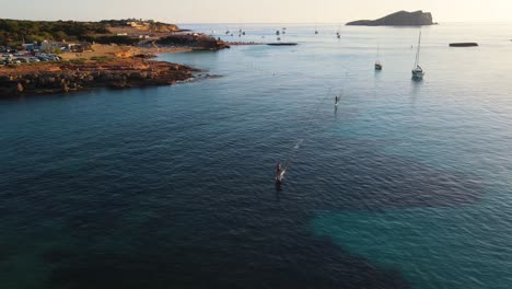 Aerial-wide-landscape-shot-of-people-surfing-with-an-eFoil-electric-surfboard-at-Cala-Escondida-in-Ibiza,-Spain