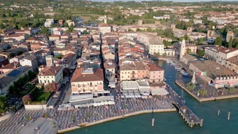 Aerial-circling-over-Lazise-at-Lake-Garda-,-promenade-with-tourists-and-constructions-of-the-town-in-the-background