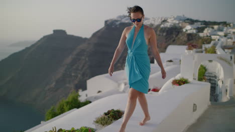 A-woman-in-a-blue-dress-walks-along-the-white-wall-during-sunset-with-Caldera-in-background---Slow-motion,-facing-forward,-Santorini,-Greece