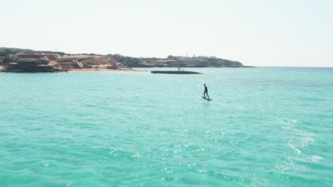 Aerial-follow-shot-of-a-man-surfing-in-an-eFoil-electric-surfboard-during-a-sunny-day-at-Cala-Escondida-in-Ibiza,-Spain
