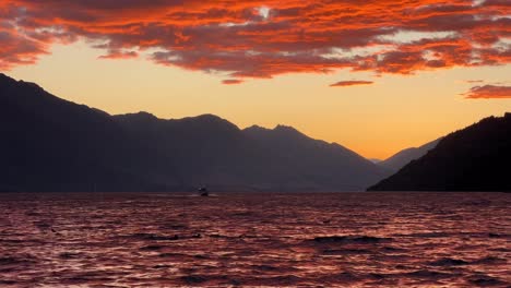 Boat-cruises-on-Lake-Wakatipu-in-Queenstown-during-golden-sunset,-orange-clouds