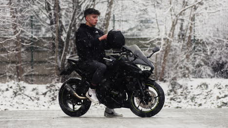 Slow-motion-of-a-young-man-wearing-his-helmet-sitting-on-a-black-Kawasaki-Ninja-650-motorcycle,-with-snow-falling-down