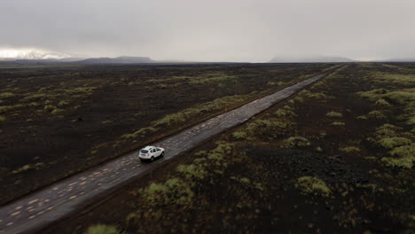 Side-aerial-follow-shot-of-white-car-driving-on-black-gravel-road-amongst-green-moss-fields-in-highlands-of-Iceland