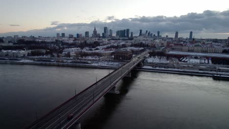 Drone-shot-showing-Vistula-River-and-driving-cars-on-bridge-in-direction-Warsaw-during-sunset-time-in-winter---Skyline-with-high-rise-buildings-in-background