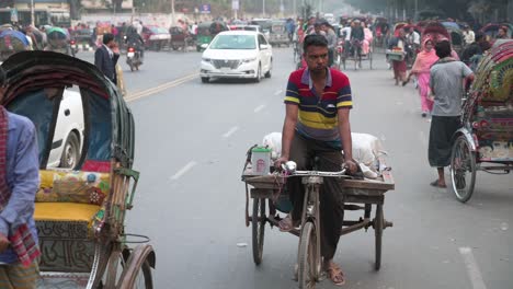 Imágenes-En-Cámara-Lenta-De-Viajeros-De-Bangladesh-Que-Viajan-En-Rickshaws-En-Una-Calle-Concurrida-Con-Otros-Vehículos-Mientras-Un-Peatón-Cruza-La-Calle-En-El-Fondo