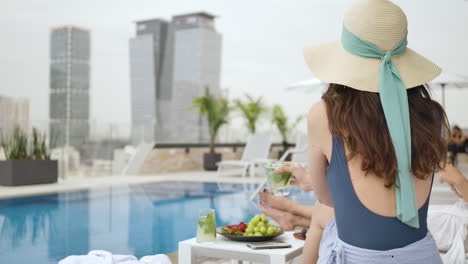 Back-View-Of-Gorgeous-Woman-In-Stylish-Summer-Hat-Sitting-Next-To-A-Pool-In-A-Hotel-Building-Rooftop