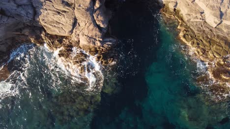 Top-down-aerial-of-cliff,-beautiful-sea-and-waves-crashing-on-rocks,-day