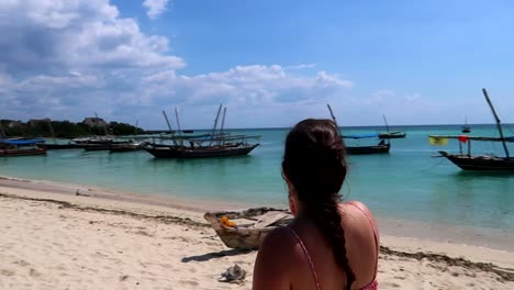 Young-woman-putting-on-sunscreen-in-slow-motion-on-the-shore-of-a-tropical-beach-with-rural-boats-docked