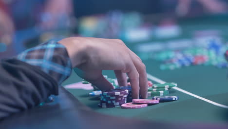 Unidentified-Person's-Hand-Holding-Colorful-Chips-On-Green-Table-With-Defocused-Background-Inside-The-Casino