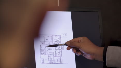 Hand-With-A-Pen-Pointing-Details-On-Floor-Plan-Room-Layout-And-Design-During-Presentation