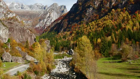 Aerial-shot-of-an-autumn-landscape-with-yellow,-green-and-red-trees,-a-rocky-river-and-mountains-on-the-horizon,-in-Val-di-Mello-in-Lombardy,-Italy