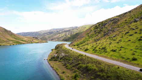 Tiny-cars-are-moving-on-a-road,-that-is-winding-between-the-mountain-and-the-calm-surface-of-a-fiord,-New-Zealand