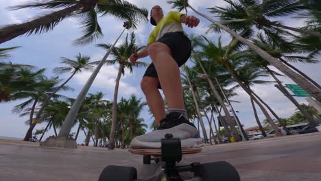 Ultra-low-angle-shot-of-a-male-skateboarder-riding-along-the-beach-using-a-longboard-with-the-view-of-tropical-palm-trees-behind-him