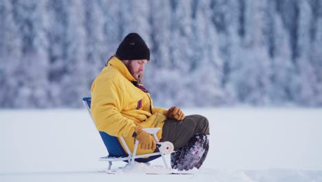 Side-Portrait-Of-A-Man-Sitting-In-Portable-Folding-Fishing-Chair,-Catching-Fish-In-Ice-Hole-With-Ice-Fishing-Rod-In-Winter
