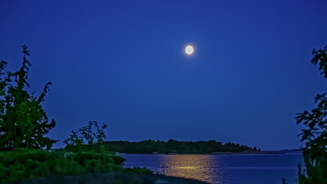 Full-moon-crossing-the-sky-over-an-island-across-a-bay---nighttime-time-lapse