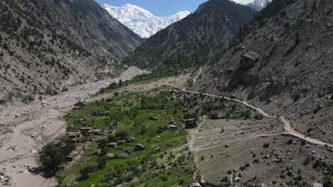 Landscape-of-Northern-Pakistan,-Aerial-View-of-Village-and-Dirt-Road-Under-Rocky-Hills-and-Snow-Capped-Mountain-Peaks