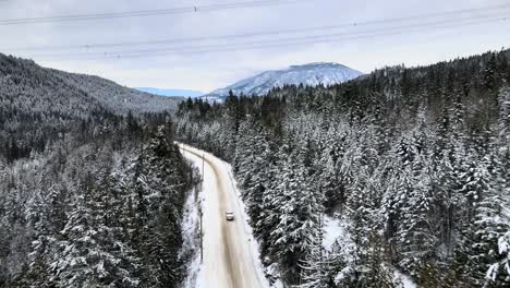 Pick-Up-Truck-on-Snow-Covered-Road-surrounded-by-Snowy-Evergreen-Forests-with-Snowy-Mountains-in-View,-drone-follow-shot