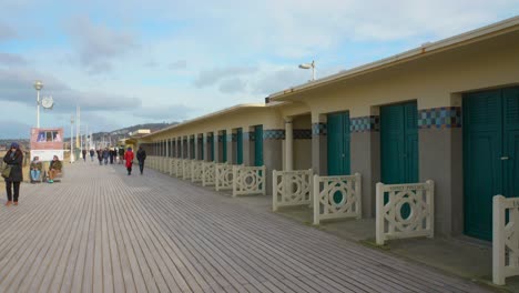 People-Walking-On-Wooden-Alley-Next-To-The-Famous-Norman-Beach-Huts-And-Wooden-Promenade-Les-Planches-Painted-With-Names-Of-American-Film-Actors-And-Directors-In-Deauville,-France