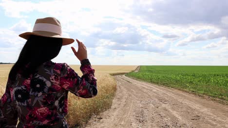 Girl-In-Dress-And-Hat-Walking-On-Dirt-Road-Through-Fields---medium-shot