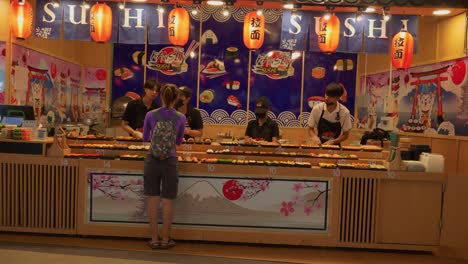 Asian-woman-standing-at-sushi-counter-as-chefs-make-and-roll-fresh-sushi-for-customers,-Thailand