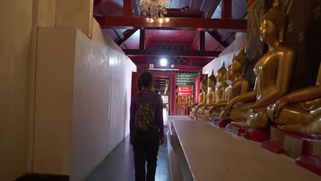 Tourists-walking-through-Buddhist-temple-passing-by-Meditation-Buddha-statues-in-a-row,-Thailand