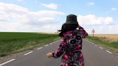 Vista-Trasera-De-Una-Mujer-Con-Vestido-Floral-Y-Sombrero-Caminando-Por-Una-Carretera-Asfaltada-Remota