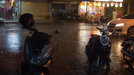 Calle-Concurrida-En-Tailandia-Con-Lluvia-Cayendo-Mientras-El-Tráfico-Pasa-Por-Un-Restaurante-Abierto-Por-La-Noche