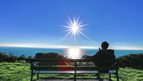 Young-man-on-a-park-bench-watching-the-ocean-with-the-sun-reflecting-off-the-water---siding-time-lapse