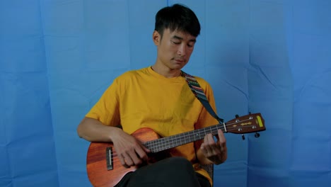 Asian-man-playing-a-stringed-ukulele-musical-instrument-with-blue-background