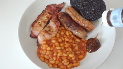 Brown-sauce-being-added-to-a-traditional-English-breakfast-of-cumberland-sausages,-back-bacon-black-pudding-and-baked-beans,-but-no-egg,-on-a-white-plate