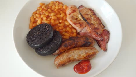 Tomato-ketchup-being-added-to-a-traditional-English-breakfast-of-cumberland-sausages,-back-bacon-black-pudding-and-baked-beans,-but-no-egg,-on-a-white-plate