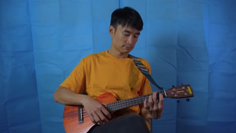 Man-slowly-playing-stringed-ukulele-musical-instrument-with-blue-background-while-seated
