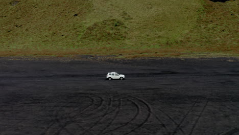 Luftaufnahme:-Seitliches-Verfolgen-Eines-Weißen-Autos,-Das-Am-Schwarzen-Sandstrand-In-Der-Nähe-Von-Grünem-Moos-In-Island-Fährt