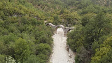 Drone-Video-of-Old-stone-three-Arch-Bridge-of-Plakidas-Zagori-region-Greece-in-forest-,-dry-river-beneath-and-rock-formation-in-the-back-panning-left-
