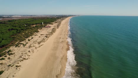 Establishing-aerial-view-over-Huelva-Spanish-golden-sandy-woodland-beach-and-coastal-lighthouse-tower-in-the-distance