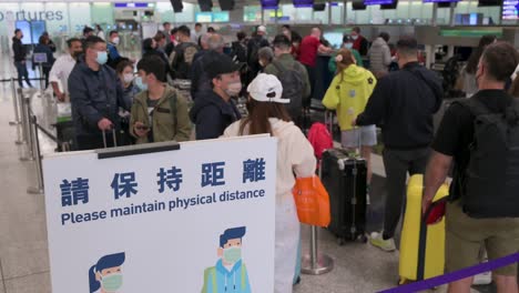Chinesische-Reisepassagiere-Warten-Am-Check-in-Schalter-Einer-Fluggesellschaft,-Während-Ein-Schild-Die-Menschen-Daran-Erinnert,-Auf-Dem-Internationalen-Flughafen-Chek-Lap-Kok-In-Hongkong-Soziale-Distanz-Zu-Wahren