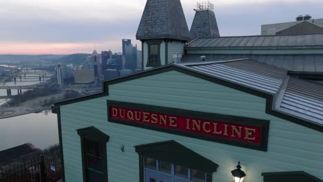 Aerial-rising-shot-of-Duquesne-Incline-building-on-top-of-Mt-Washington-with-Pittsburgh,-Pennsylvania-skyline-and-three-rivers-in-background-at-sunrise