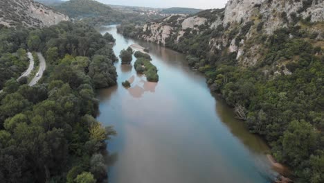 Drohnenvideo-Fliegt-über-Den-Fluss-Nestos-In-Griechenland,-Felsen-Und-Klippen-Auf-Der-Rechten-Seite-Und-Bäume-Und-Büsche-Auf-Der-Linken-Seite,-Ein-Dorf-In-Der-Ferne-An-Einem-Sommertag